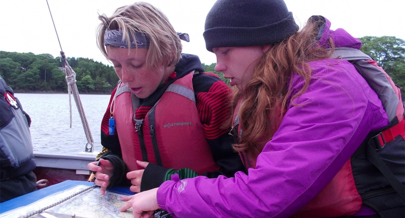 Two young people wearing lifejackets examine a map while sitting on a sailboat. 
